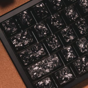 May include: A mechanical keyboard featuring custom keycaps with a black and gray forged carbon fiber pattern. The keys display standard white lettering including ESC, TAB, CAPS, SHIFT, and alphanumeric characters arranged in a standard layout.