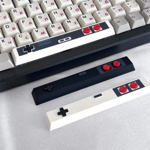 Peut inclure: Trois barres d'espace pour clavier mécanique arborant des designs de manettes de jeu vidéo rétro. L'une est installée sur un clavier aux touches blanches avec des caractères cyrilliques, deux autres, une noire et une blanche, sont posées dessous.
