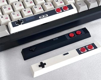 May include: Three mechanical keyboard spacebars featuring retro video game controller designs. One is installed on a keyboard with white keys and Cyrillic characters, while two separate bars, one black and one white, are displayed below it.