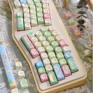 K&ouml;nnte beinhalten: Eine pastellfarbene Tastatur mit individuellen Tastenkappen. Die Tastenkappen zeigen verschiedene Illustrationen und Symbole in Rosa, Gr&uuml;n, Blau und Gelb. Die Tastatur hat eine cremefarbene Basis und ein einzigartiges, k&uuml;nstlerisches Design.