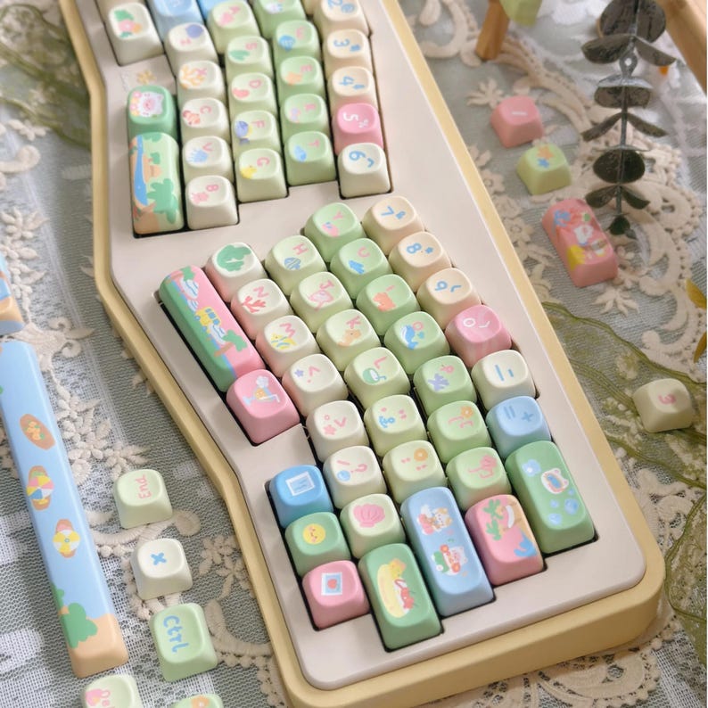 K&ouml;nnte beinhalten: Eine pastellfarbene Tastatur mit individuellen Tastenkappen. Die Tastenkappen zeigen verschiedene Illustrationen und Symbole in Rosa, Gr&uuml;n, Blau und Gelb. Die Tastatur hat eine cremefarbene Basis und ein einzigartiges, k&uuml;nstlerisches Design.