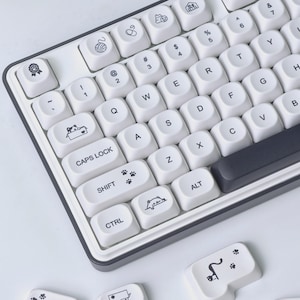Puede incluir: Un teclado mecánico con teclas blancas, letras negras e iconos de gatos, incluyendo huellas, un gato en una caja y un gato asomándose por el borde. El teclado tiene un marco y una barra espaciadora de color gris oscuro.