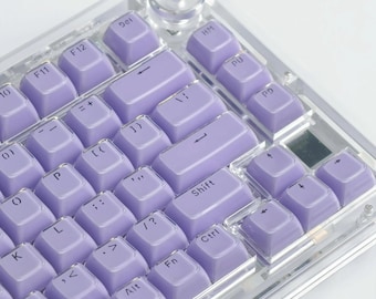 Könnte beinhalten: Eine mechanische Tastatur mit einem transparenten Rahmen und hellvioletten Tastenkappen. Die Tastenkappen haben weiße Buchstaben und Symbole. Die Tastatur hat einen Lautstärkeregler und verschiedene Funktionstasten.