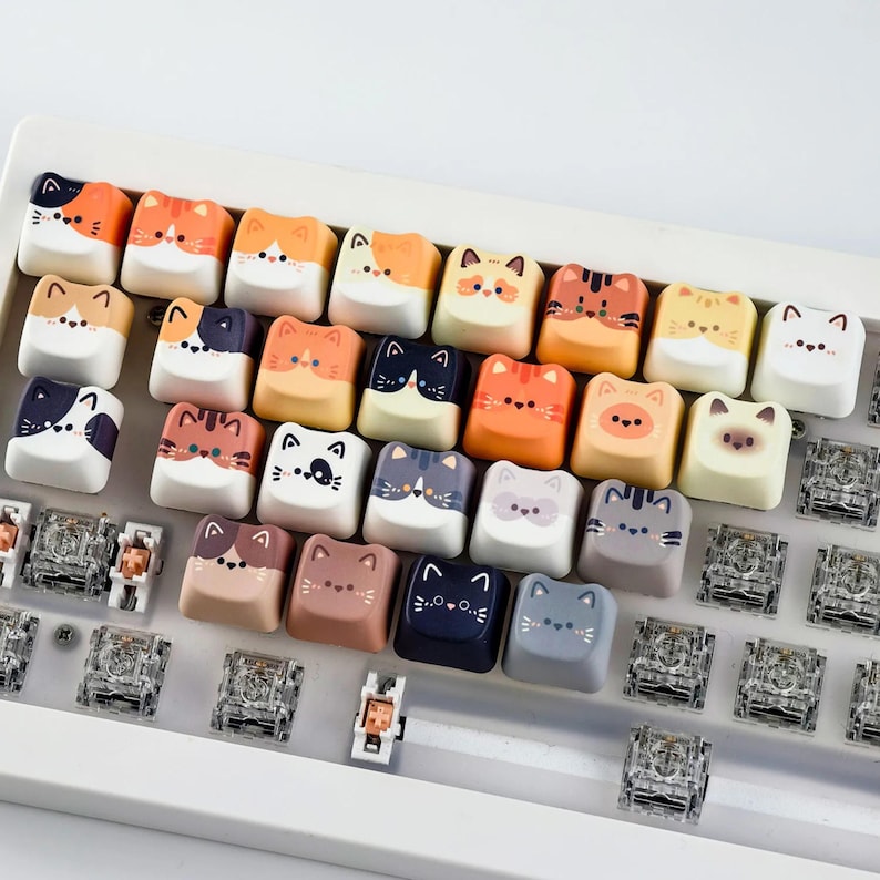 K&ouml;nnte beinhalten: Eine Sammlung von Tastenkappen im Katzen-Design in verschiedenen Farben, darunter Orange, Wei&szlig;, Schwarz und Grau. Jede Tastenkappe zeigt ein anderes Katzengesicht-Design. Die Tastenkappen sind auf einer wei&szlig;en Tastatur angeordnet und zeigen eine verspielte und einzigartige &Auml;sthetik.