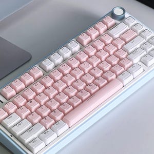 May include: A pastel-colored mechanical keyboard with a light blue frame. The keycaps alternate between white and pink, with a pink space bar. A silver knob is in the upper right corner. The keyboard is on a light gray surface.