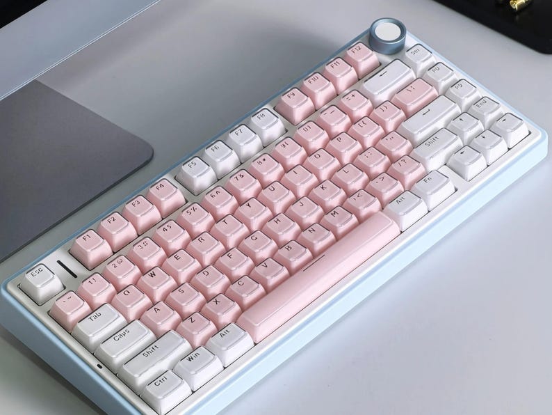 May include: A pastel-colored mechanical keyboard with a light blue frame. The keycaps alternate between white and pink, with a pink space bar. A silver knob is in the upper right corner. The keyboard is on a light gray surface.