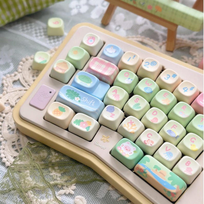 K&ouml;nnte beinhalten: Eine pastellfarbene Tastatur mit individuellen Tastenkappen. Die Tasten zeigen verschiedene Designs, darunter Fr&uuml;chte, Blumen und Tiere. Die "Shift"-Taste ist hellblau, und die Tastatur hat einen hellgelben Rahmen. Das Design ist niedlich und verspielt.