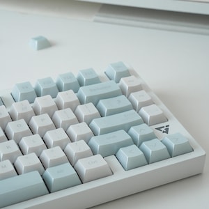 Könnte beinhalten: Eine mechanische Tastatur mit hellblauen und weißen Tastenkappen. Das Layout umfasst standardmäßige alphanumerische Tasten, eine Rücktaste, eine Eingabetaste und Pfeiltasten. Auf dem Tastaturrahmen ist ein kleines dreieckiges Logo aufgedruckt.