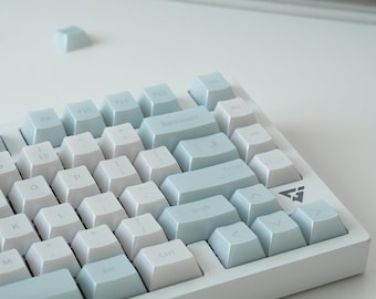 Peut inclure: Un clavier mécanique doté d'un jeu de touches bleu clair et blanc. La disposition comprend des touches alphanumériques standard, une touche retour arrière, une touche entrée et des touches fléchées. Un petit logo triangulaire est imprimé.