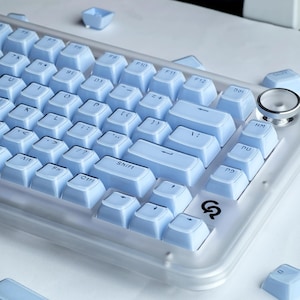 Puede incluir: Un teclado mec&aacute;nico azul claro con letras blancas. El teclado tiene un marco de pl&aacute;stico transparente y teclas azules claras. Las teclas incluyen letras, n&uacute;meros y teclas de funci&oacute;n. El teclado est&aacute; dise&ntilde;ado para juegos o escribir.