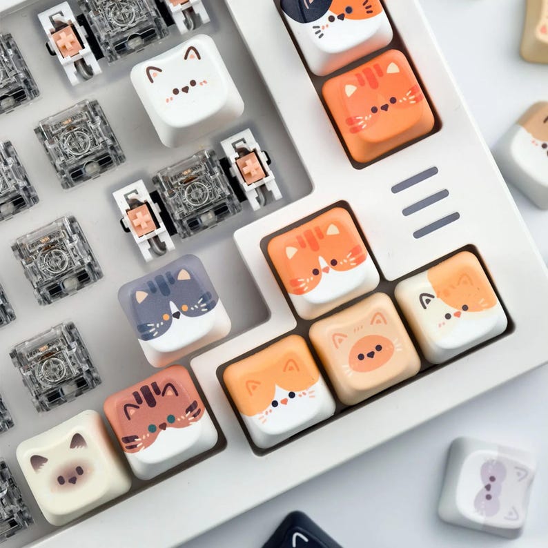 K&ouml;nnte beinhalten: Nahaufnahme einer wei&szlig;en Tastatur mit individuellen Tastenkappen mit Katzenmotiven in verschiedenen Farben. Die Tastenkappen sind orange, wei&szlig;, grau und beige. Transparente mechanische Tastaturschalter sind ebenfalls sichtbar.