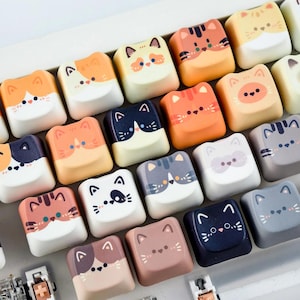 K&ouml;nnte beinhalten: Eine Sammlung farbenfroher Tastaturkappen mit entz&uuml;ckenden Katzenmotiven. Die Kappen zeigen verschiedene Katzenrassen mit unterschiedlichen Fellmustern und -farben, darunter Orange, Grau und Schildpatt. Die quadratischen Kappen haben eine glatte Oberfl&auml;che.