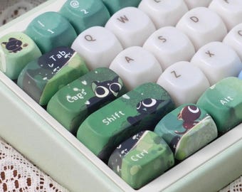 Könnte beinhalten: Eine individuelle Tastatur mit grünen, weißen und türkisfarbenen Tastenkappen. Einige Tastenkappen zeigen Katzenillustrationen und Landschaftsdesigns. Die Tasten umfassen "Tab", "Caps", "Shift" und "Ctrl".