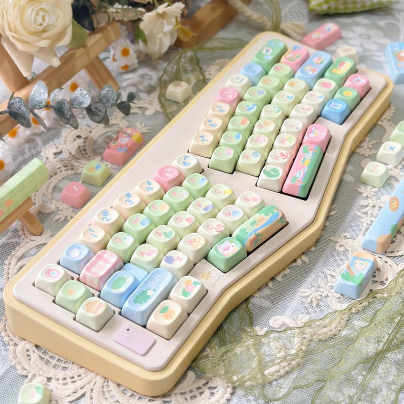 K&ouml;nnte beinhalten: Eine pastellfarbene mechanische Tastatur mit individuellen Tastenkappen. Die Tastenkappen weisen verschiedene Designs auf, darunter Fr&uuml;chte, Tiere und Muster in Rosa-, Gr&uuml;n-, Blau- und Gelbt&ouml;nen. Die Tastatur hat eine cremefarbene Basis und eine einzigartige, ergonomische Form.
