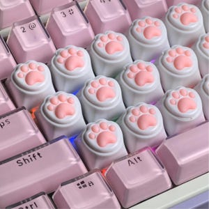 May include: A mechanical keyboard featuring light pink keycaps and several white custom keycaps shaped like cat paws with raised pink pads. The keys include standard alphanumeric characters, a Shift key, and an Alt key with black lettering.