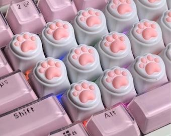 May include: A mechanical keyboard featuring light pink keycaps and several white custom keycaps shaped like cat paws with raised pink pads. The keys include standard alphanumeric characters, a Shift key, and an Alt key with black lettering.