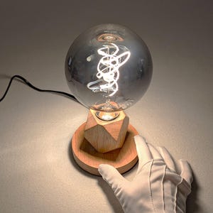 Peut inclure: Une lampe de table décorative avec une grande ampoule grise en forme de globe. L'ampoule présente un design de filament tourbillonnant unique. La lampe repose sur une base en bois au design géométrique.