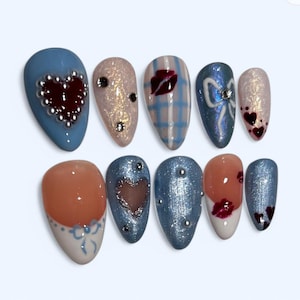 Puede incluir: Un conjunto de diez uñas postizas en forma de almendra. Los diseños incluyen una uña azul con un corazón de cuentas plateadas, una uña rosa con purpurina y pedrería, y una uña a cuadros azules con un diseño de labios rojos. Otras uñas presentan lazos, corazones y purpurina.