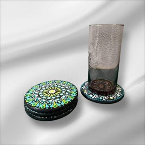 Könnte beinhalten: Ein Satz schwarzer Untersetzer mit einem farbenfrohen Mandala-Design. Die Untersetzer weisen ein Muster aus teal, gelben und weißen Punkten und tropfenförmigen Formen auf. Ein hohes Glas steht auf einem der Untersetzer.