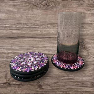 Könnte beinhalten: Ein Set aus vier schwarzen Untersetzern mit einem Mandala-Design in Lila-, Rosa- und Weißtönen. Ein hohes Glas steht auf einem passenden Untersetzer. Die Untersetzer sind auf einer Holzoberfläche gestapelt.