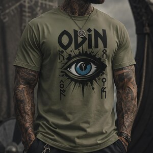 May include: Olive green t-shirt with the word "ODIN" and an eye graphic. The eye is blue with black rays. The person is wearing a silver necklace and black pants. A Viking ship is in the background.