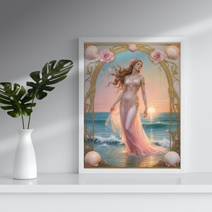 Op de afbeelding: Een ingelijste kunstdruk toont een vrouw in een roze jurk in de zee bij zonsondergang. Het kunstwerk heeft een gouden boog, schelpen en rozen. Een witte vaas met groene bladeren staat op een witte plank.