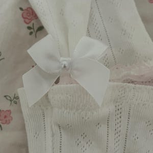 May include: Close-up of white, open-knit fabric with a delicate pattern. A white satin bow with a pearl bead center adorns the fabric. The background features a floral pattern in pink and green.