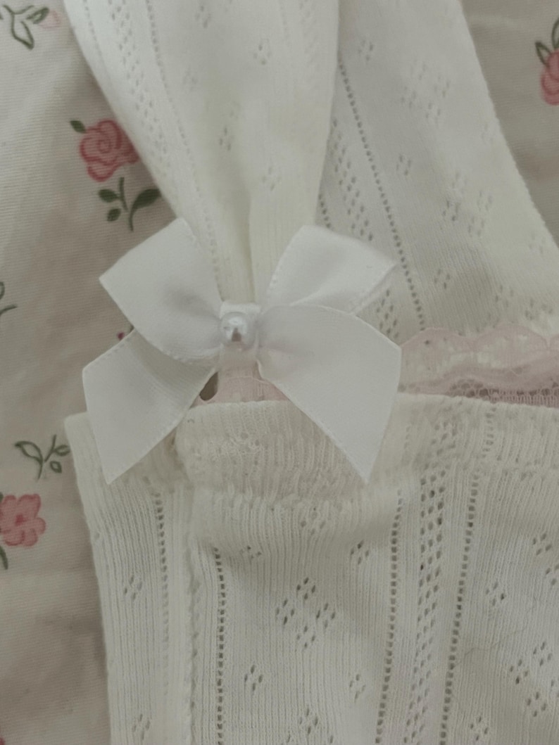 May include: Close-up of white, open-knit fabric with a delicate pattern. A white satin bow with a pearl bead center adorns the fabric. The background features a floral pattern in pink and green.