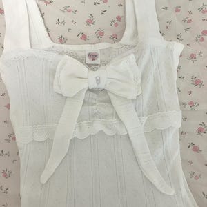 May include: White camisole with a large bow and lace trim. The camisole has a textured pattern and a small tag that reads "Couse". The bow is centered on the chest and has a small jewel in the center.