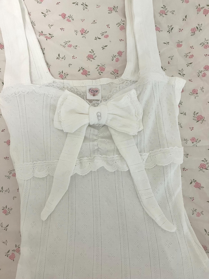 May include: White camisole with a large bow and lace trim. The camisole has a textured pattern and a small tag that reads "Couse". The bow is centered on the chest and has a small jewel in the center.