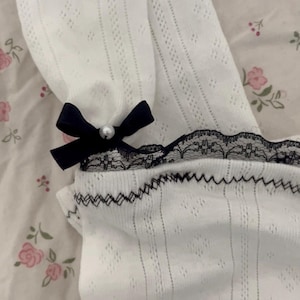 May include: White, open-knit fabric with a black lace trim and a small black bow with a pearl accent. The fabric has a delicate, airy appearance. The lace trim is detailed with small pearls. The bow adds a touch of elegance.