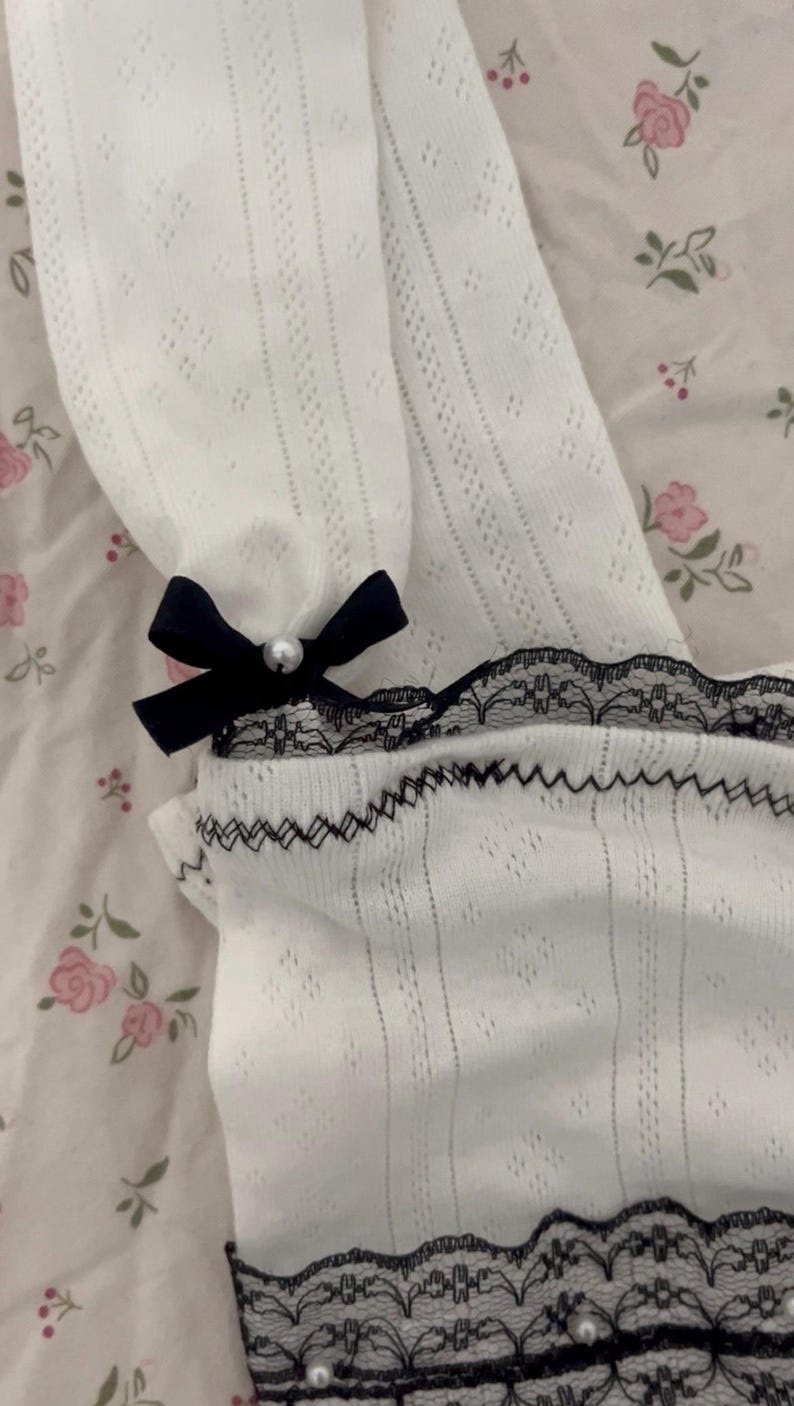 May include: White, open-knit fabric with a black lace trim and a small black bow with a pearl accent. The fabric has a delicate, airy appearance. The lace trim is detailed with small pearls. The bow adds a touch of elegance.