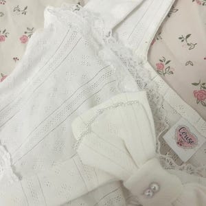 May include: White, eyelet-detailed camisole with lace trim and a bow accent. The bow is adorned with small pearls. The camisole is made of a soft, textured fabric. The tag reads "Corise".