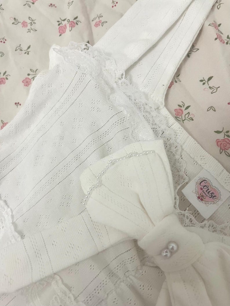 May include: White, eyelet-detailed camisole with lace trim and a bow accent. The bow is adorned with small pearls. The camisole is made of a soft, textured fabric. The tag reads "Corise".