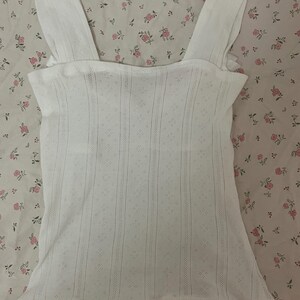May include: White eyelet tank top with wide shoulder straps. The top features a textured, open-weave design with vertical and diamond patterns. The garment is made of a lightweight fabric, suitable for warm weather.