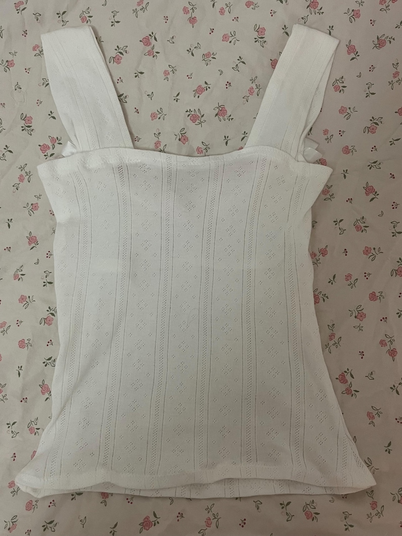 May include: White eyelet tank top with wide shoulder straps. The top features a textured, open-weave design with vertical and diamond patterns. The garment is made of a lightweight fabric, suitable for warm weather.