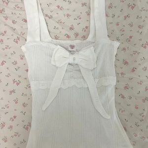 May include: White tank top with a large bow and lace trim. The top has a textured, vertical striped pattern and wide straps. The bow is centered on the front, and the top is displayed on a floral patterned fabric.