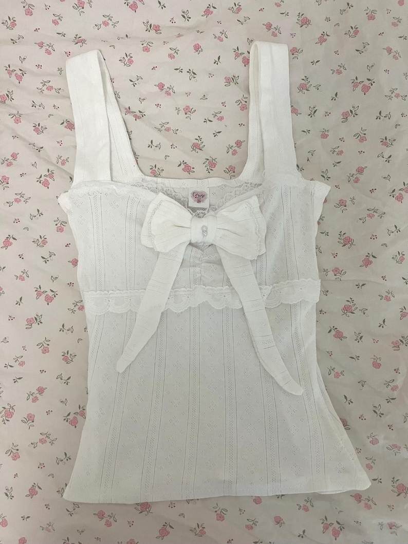 May include: White tank top with a large bow and lace trim. The top has a textured, vertical striped pattern and wide straps. The bow is centered on the front, and the top is displayed on a floral patterned fabric.