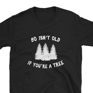 Op de afbeelding: Zwart T-shirt met witte tekst die "80 isn't old if you're a tree" luidt. De tekst wordt omringd door een afbeelding van drie gestileerde bomen.