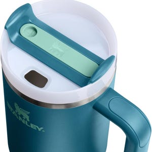 K&ouml;nnte beinhalten: Nahaufnahme eines teal-farbenen Stanley-Reisebechers mit wei&szlig;em Rand und teal-farbenem Griff. Der Deckel hat einen teal-farbenen und mintgr&uuml;nen Schieber mit dem Stanley-Logo. Geeignet f&uuml;r hei&szlig;e oder kalte Getr&auml;nke.