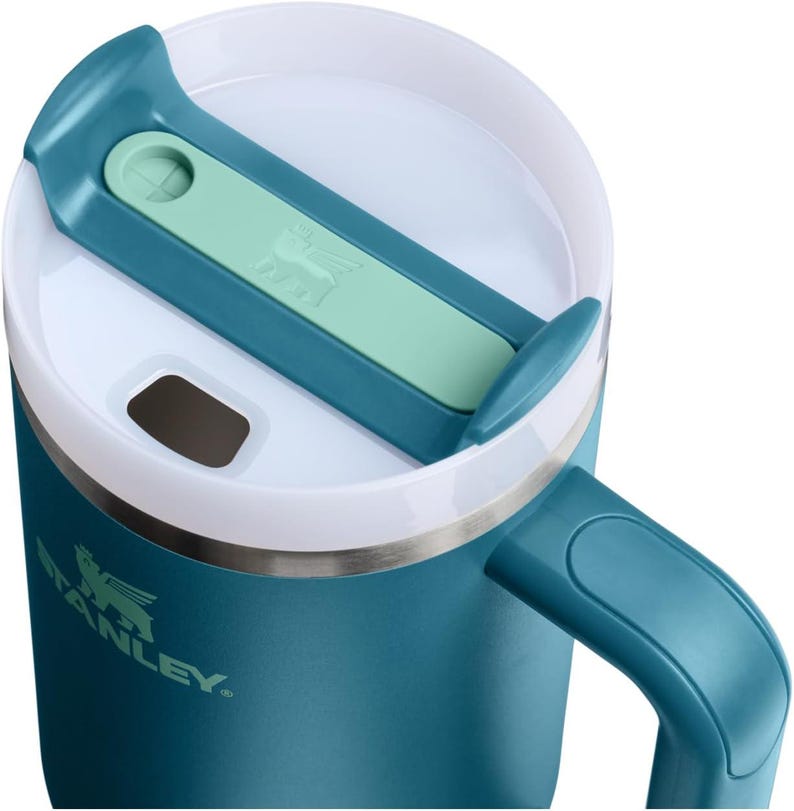 K&ouml;nnte beinhalten: Nahaufnahme eines teal-farbenen Stanley-Reisebechers mit wei&szlig;em Rand und teal-farbenem Griff. Der Deckel hat einen teal-farbenen und mintgr&uuml;nen Schieber mit dem Stanley-Logo. Geeignet f&uuml;r hei&szlig;e oder kalte Getr&auml;nke.