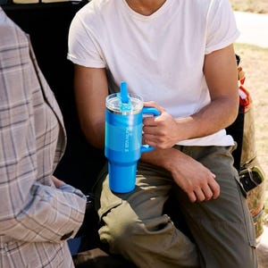 Puede incluir: Un vaso Stanley azul brillante con asa y tapa transparente. El vaso tiene un agarre texturizado y el logotipo de Stanley. La persona lleva una camiseta blanca y pantalones verde oliva.