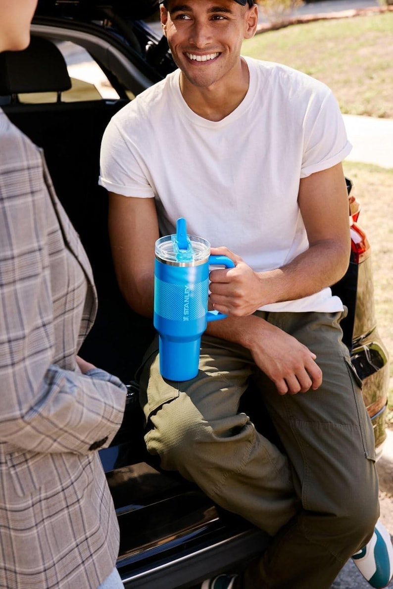 Puede incluir: Un vaso Stanley azul brillante con asa y tapa transparente. El vaso tiene un agarre texturizado y el logotipo de Stanley. La persona lleva una camiseta blanca y pantalones verde oliva.