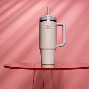 May include: A cream-colored Stanley tumbler with a handle and straw, set on a red table against a pink backdrop. The tumbler features the Stanley logo and a silver band near the top. The design is sleek and modern.