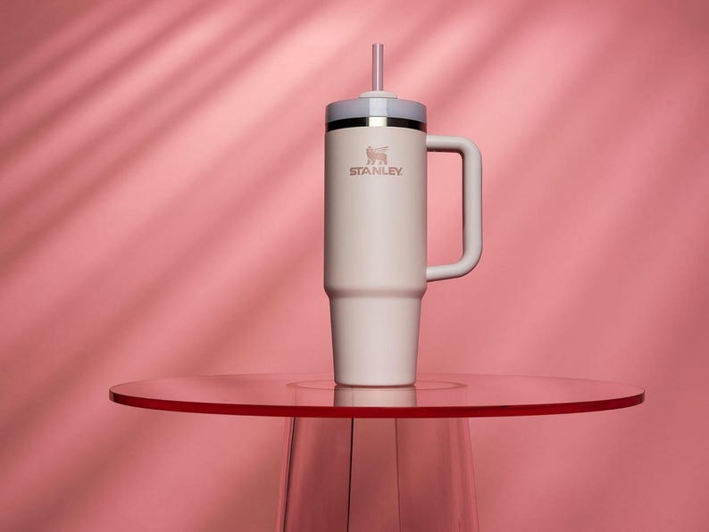 May include: A cream-colored Stanley tumbler with a handle and straw, set on a red table against a pink backdrop. The tumbler features the Stanley logo and a silver band near the top. The design is sleek and modern.