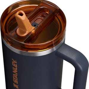 Puede incluir: Primer plano de un termo Stanley azul marino con tapa abatible y asa marrones. El termo tiene un borde plateado y el logotipo de Stanley en naranja. El asa tiene un patr&oacute;n texturizado. La tapa est&aacute; parcialmente abierta.
