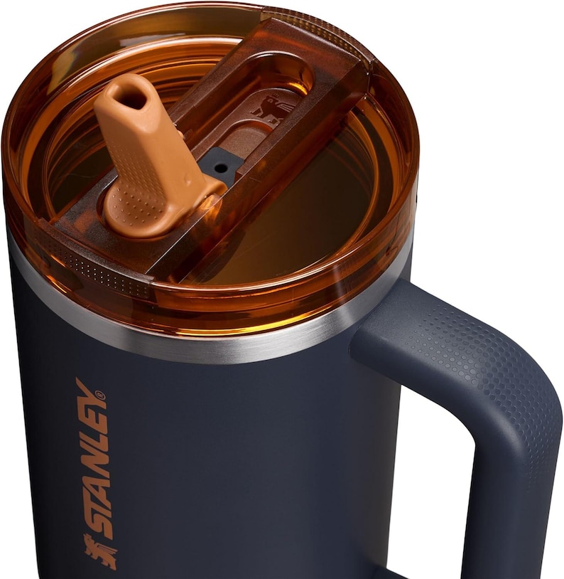 Puede incluir: Primer plano de un termo Stanley azul marino con tapa abatible y asa marrones. El termo tiene un borde plateado y el logotipo de Stanley en naranja. El asa tiene un patr&oacute;n texturizado. La tapa est&aacute; parcialmente abierta.