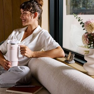 K&ouml;nnte beinhalten: Ein hellrosa isolierter Becher mit Griff und wei&szlig;em Deckel. Der Becher wird von einer Person gehalten, die ein wei&szlig;es Hemd und gestreifte Hosen tr&auml;gt. Eine Vase mit rosa Blumen steht auf einer Fensterbank.