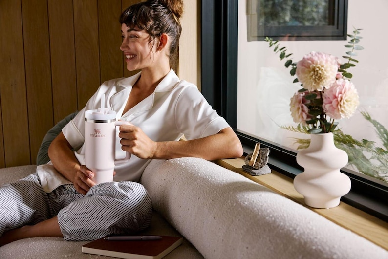 K&ouml;nnte beinhalten: Ein hellrosa isolierter Becher mit Griff und wei&szlig;em Deckel. Der Becher wird von einer Person gehalten, die ein wei&szlig;es Hemd und gestreifte Hosen tr&auml;gt. Eine Vase mit rosa Blumen steht auf einer Fensterbank.
