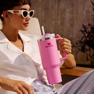 K&ouml;nnte beinhalten: Ein gro&szlig;er pinkfarbener Stanley-Becher mit Griff und Strohhalm. Der Becher hat einen wei&szlig;en Deckel und das Stanley-Logo. Die Person tr&auml;gt eine wei&szlig;e Sonnenbrille und ein wei&szlig;es Hemd.